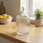 Clear glass jar with a lid on a wooden surface, with a bowl of lemons and a plant in the background.