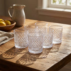 Set of six clear glass tumblers on a wooden table with a bowl of lemons and a ceramic pitcher in the background.