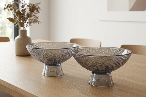 Two decorative glass bowls on a wooden table with a vase of dried flowers in the background.