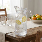 Clear glass pitcher with lemon slices on a wooden table with glasses and fruit in the background.