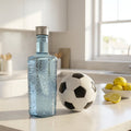 Clear glass bottle with silver cap on a kitchen counter with a soccer ball and lemons.