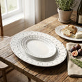 Set of white ceramic plates with decorative edge on a wooden table with food and a plant in the background.